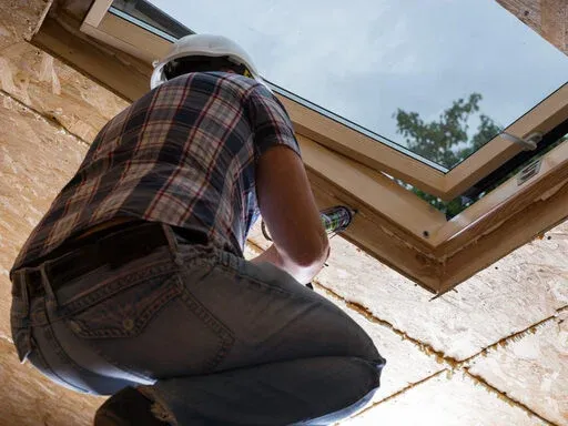 A construction worker wearing a helmet crouches to install a skylight construction worker, installing a roll of roofing material