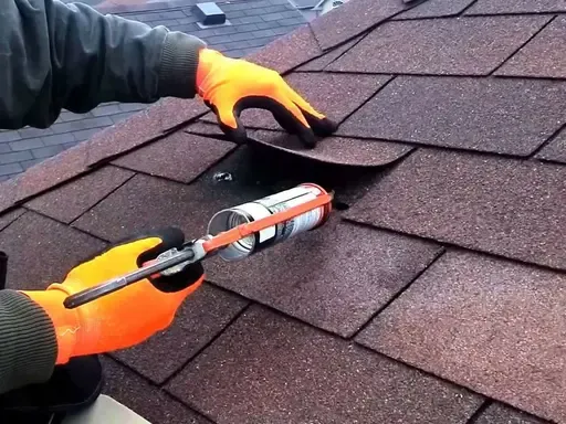 A worker applies sealant to a loose shingle on a brown asphalt roof A worker applies sealant to a loose shingle on a brown asphalt roof