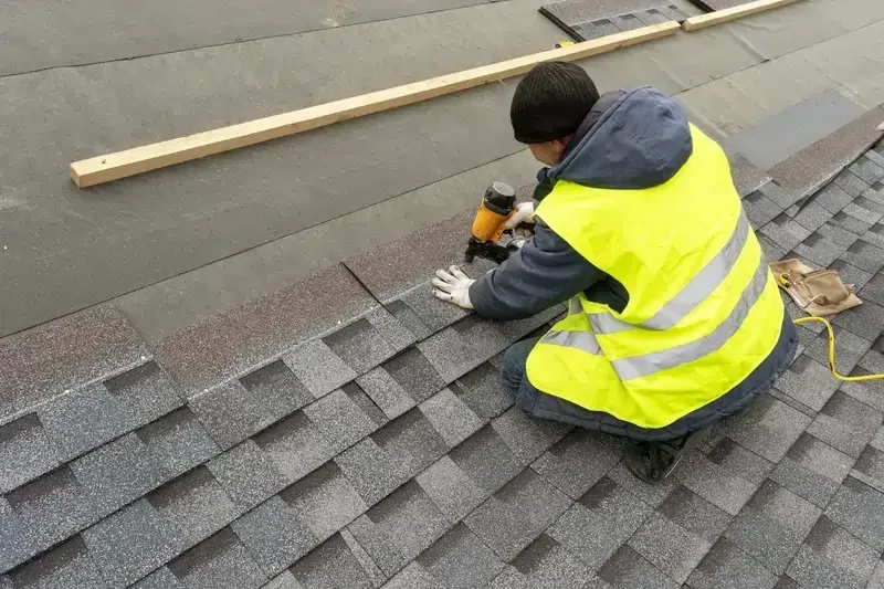 A worker in a yellow safety vest installs shingles on a roof using a nail gun A worker in a yellow safety vest installs shingles on a roof using a nail gun